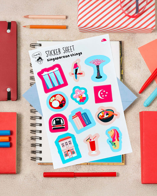 Sticker sheet with colorful Singapore-themed illustrations on a notebook surrounded by stationery items.