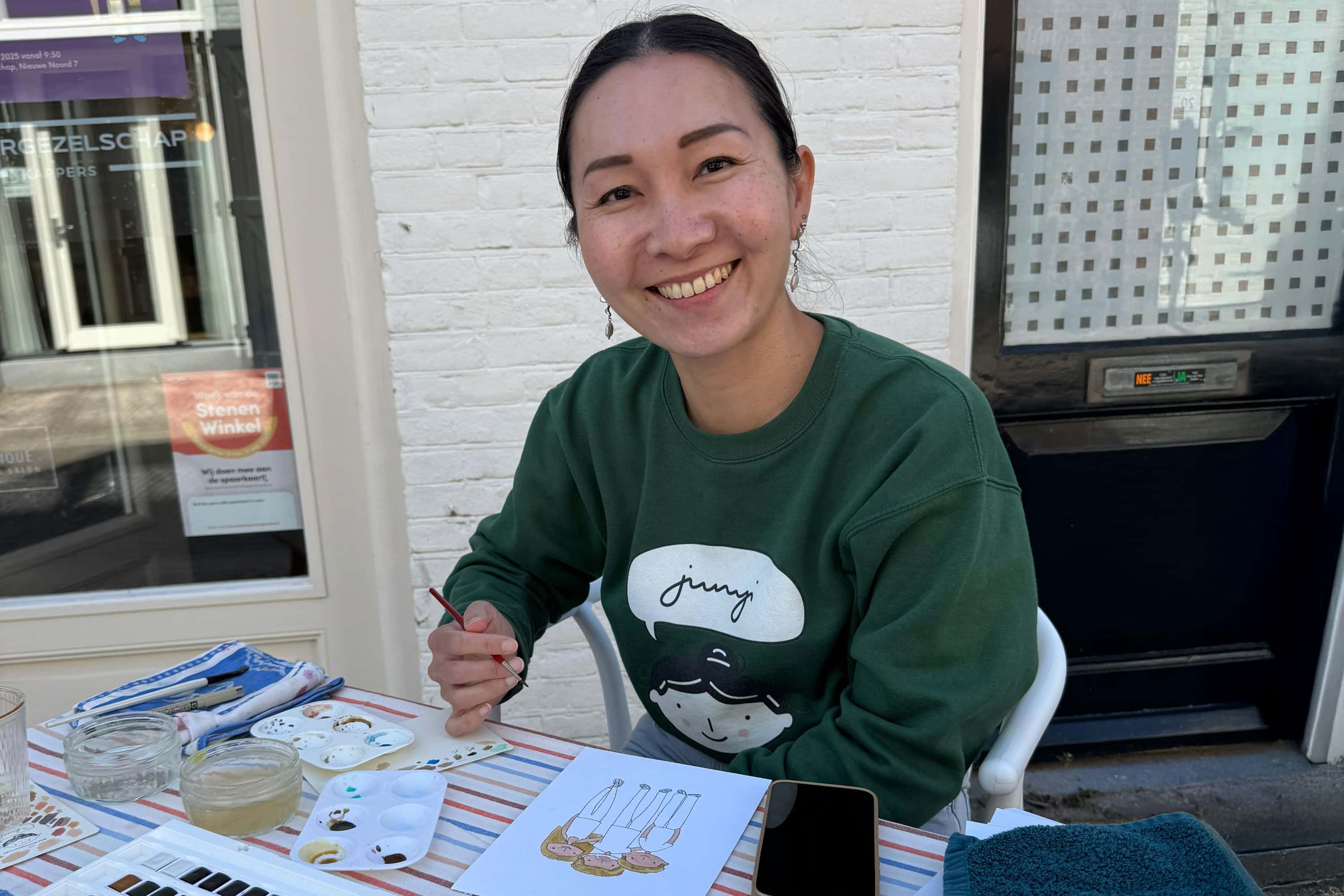 Jun-Yi sitting at a table with art supplies, smiling, during a Mother's Day live painting session.