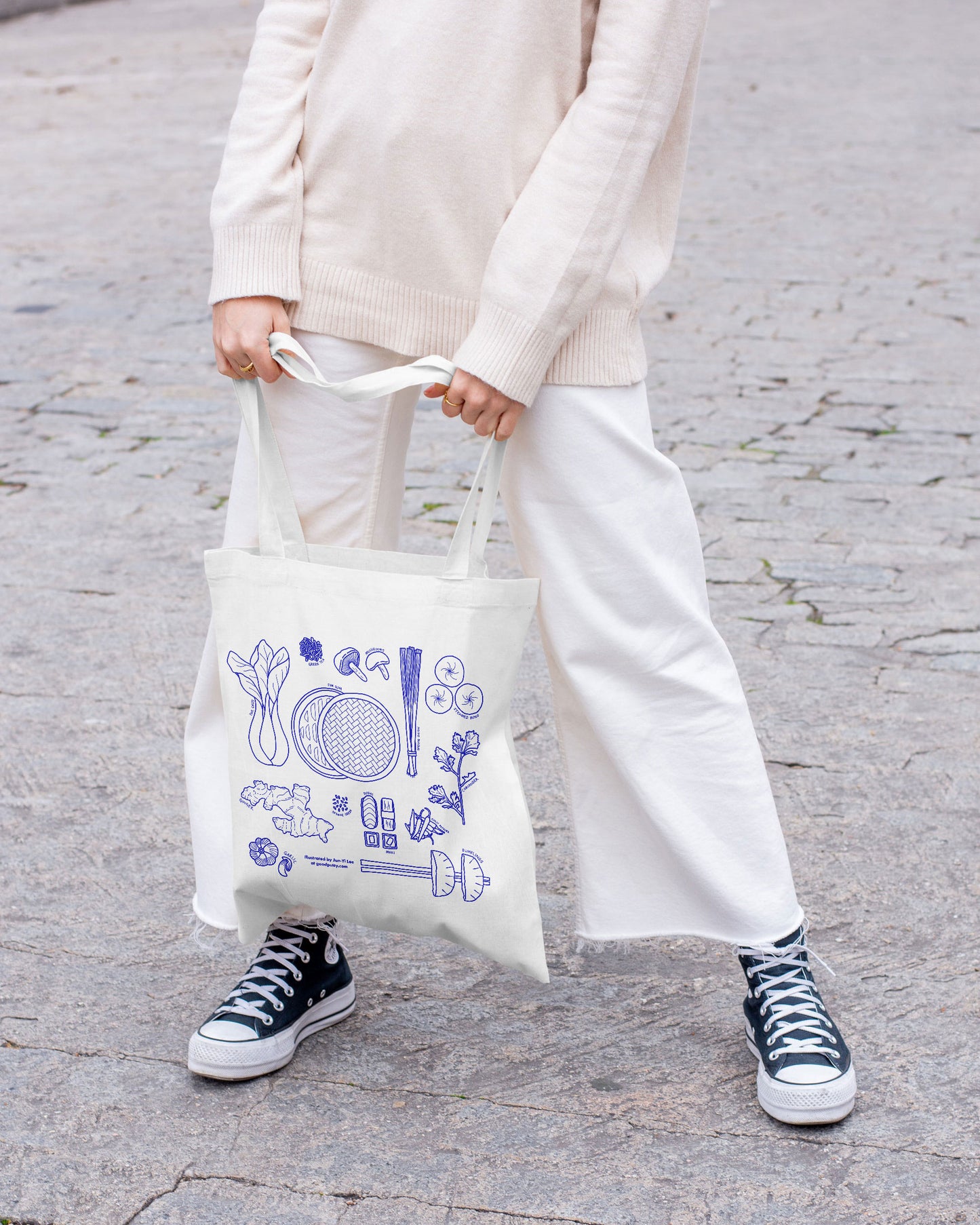 Person holding a tote bag with blue illustrations of Asian ingredients on a gray pavement background
