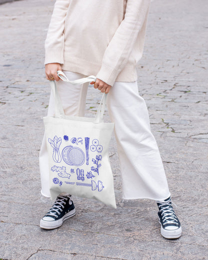 Person holding a tote bag with blue illustrations of Asian ingredients on a gray pavement background
