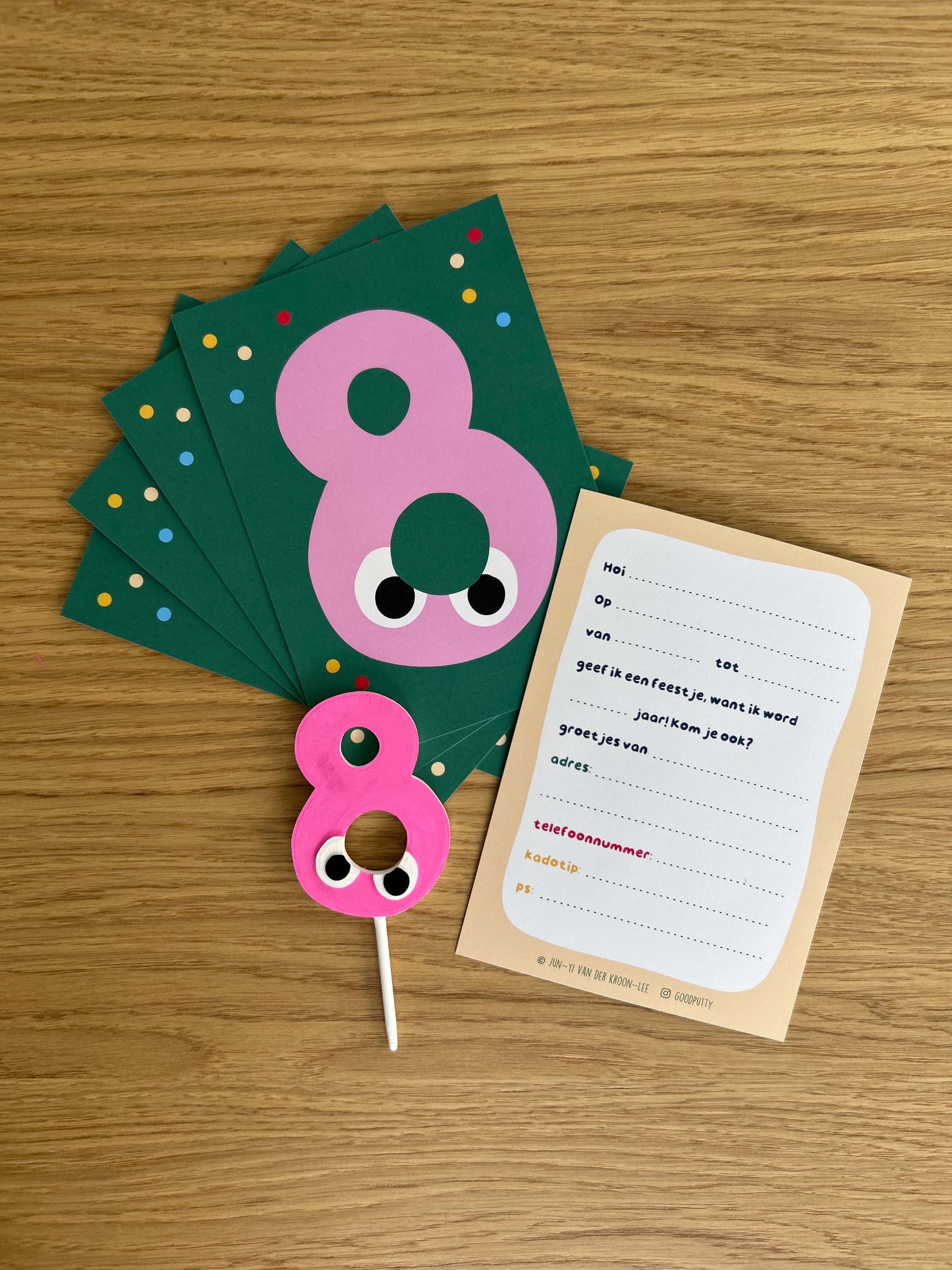 Pink number 8 cake topper with googly eyes on a wooden surface, accompanied by green cards and a paper with text.