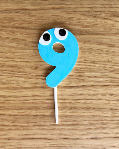 Blue number 9 cake topper with eyes on a wooden surface