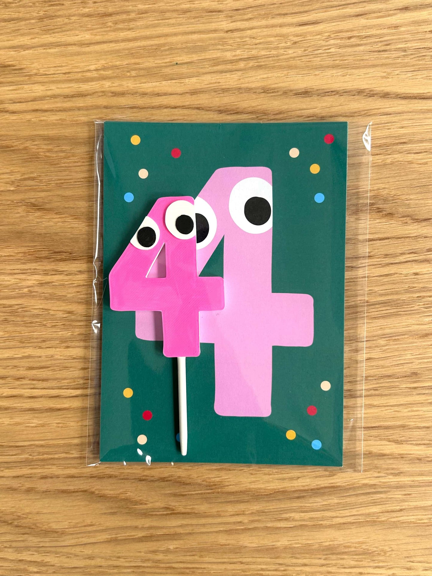 Pink number '4' with googly eyes on a green card with colorful dots, on a wooden surface.