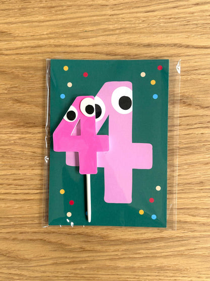 Pink number '4' with googly eyes on a green card with colorful dots, on a wooden surface.