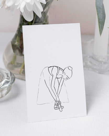 Postcard with a line drawing of a ballerina tying her shoe on a white surface with flowers in the background.