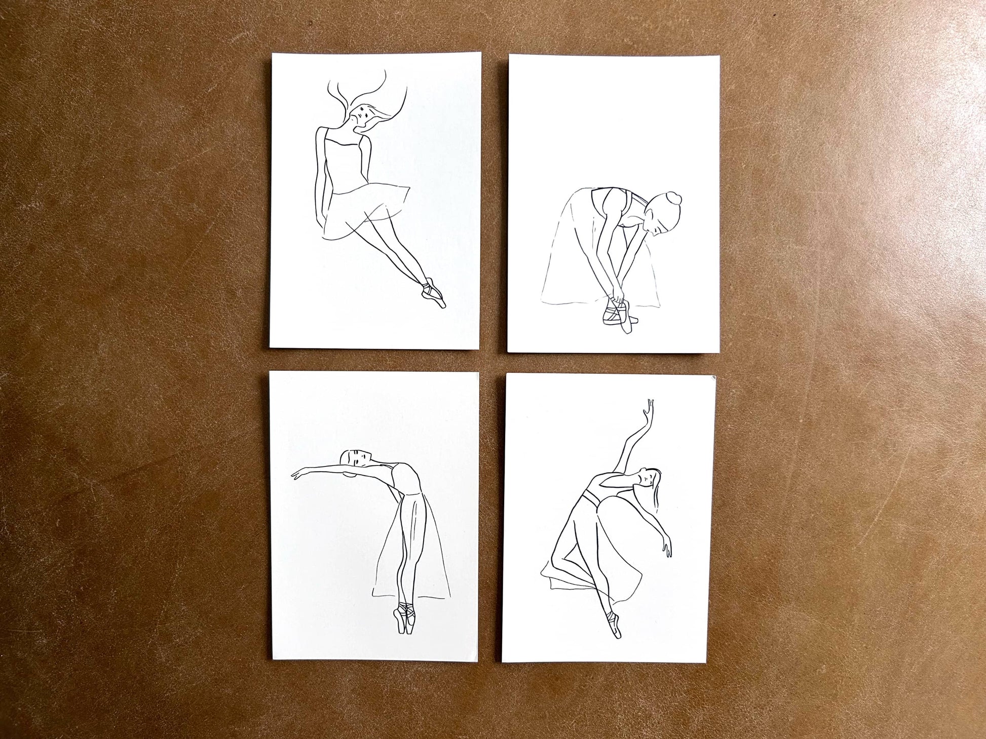 Four white cards with black line art of ballerinas on a brown leather surface