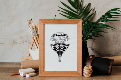Framed artwork with a hot air balloon design and a traveller's quote by J.R.R. Tolkien, placed on a wooden surface with plants and stationery items.